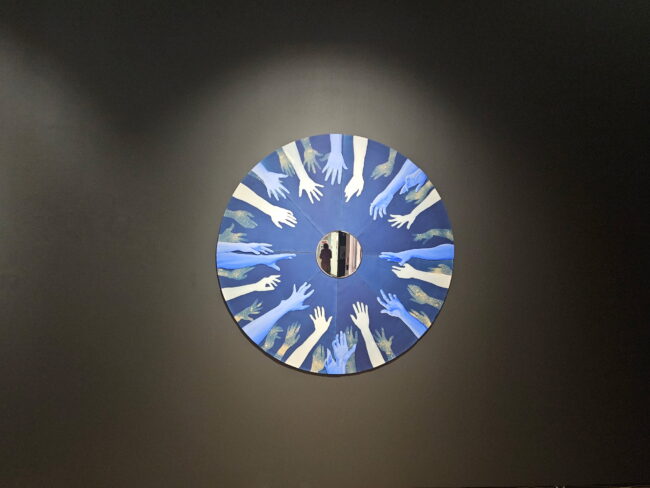 "Traces of Blue" by Lauren Tarter a circular canvas with hands reaching towards the center where a figure is shown. (Greyson Boutin/The Vista)