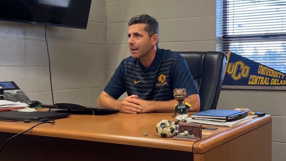 UCO Women's Soccer Coach, Amel Muhamedagic, sitting at his desk talking to someone out of frame.