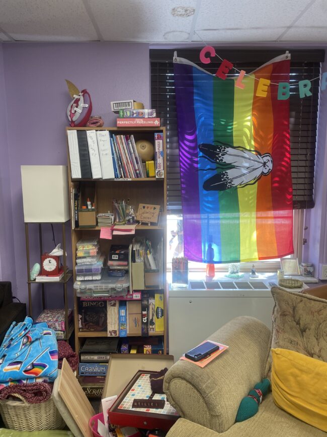 A corner of a room is cluttered with comforting items, books, lounge chairs, and games. 