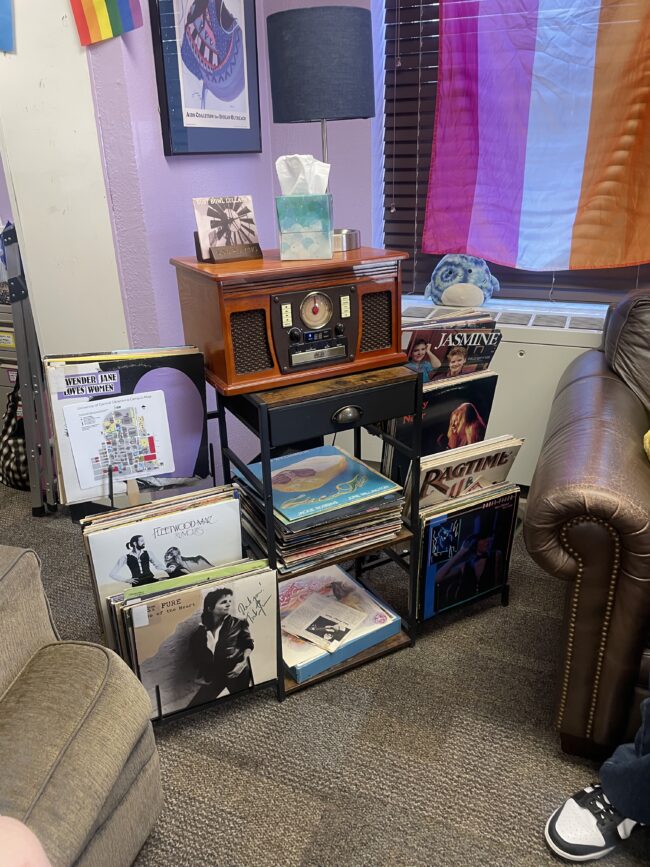 A record player is displayed on a black shelf with stacks of vinyl records around it. 