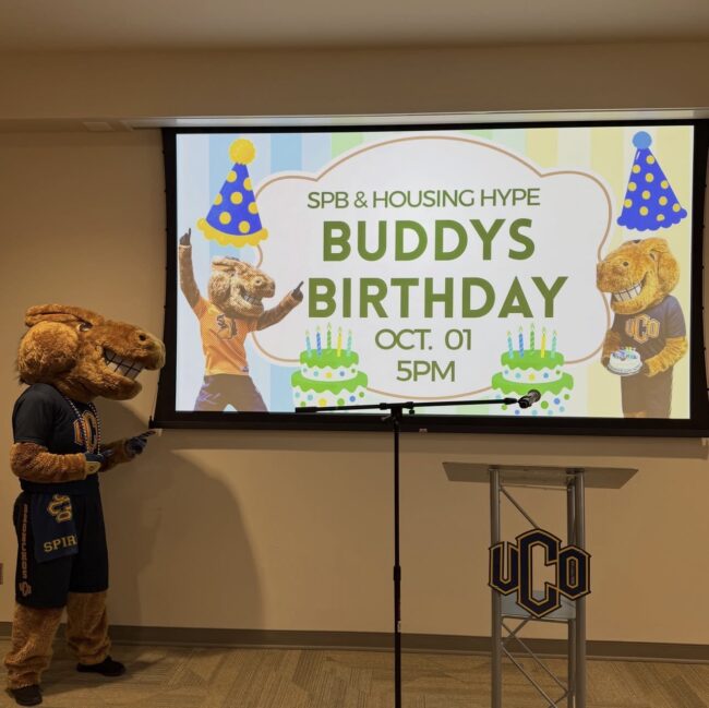 Buddy The Broncho is posing next to a projected banner reading "SPB & HOUSING HYPE. BUDDYS BIRTHDAY OCT. 1 5PM"