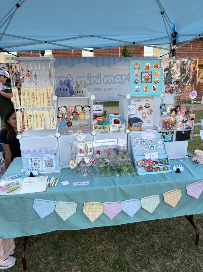 "mini mart" in bubble letters is featured on a backdrop. various keychains, plushies, stickers, and other trinkets are on display.