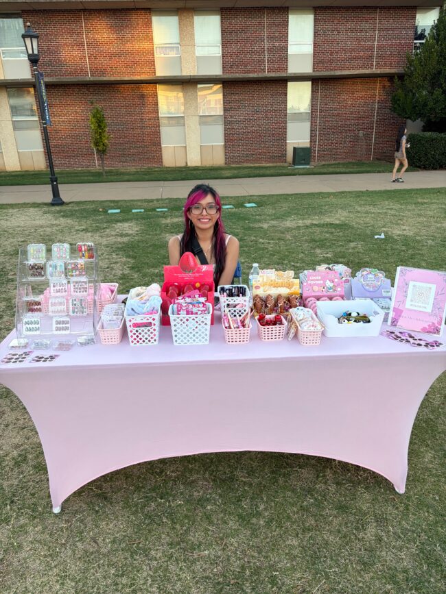 Roselyn Zoti sits at her Cosmetic Book smiling. She is selling different colored nails with various designs, and eye makeup.