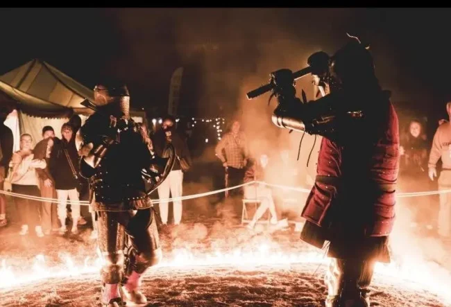 Two scare-actors dressed in full costumes are surrounded by a ring of fire, and a crowd of people as they play-fight.