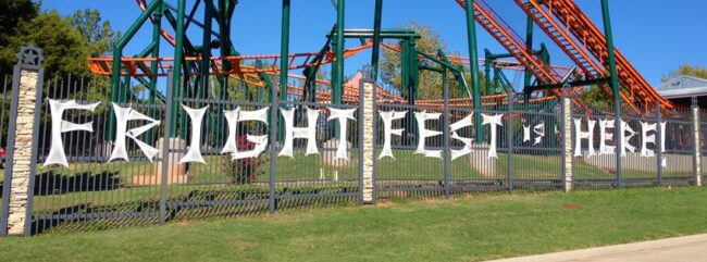 "FRIGHTFEST IS HERE!" is displayed using faux cobwebs on the fence of Frontier City. Rollercoasters can be seen in the background.